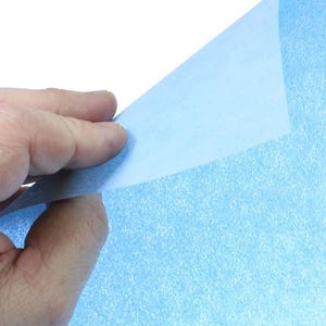 May include: A sheet of light blue glitter paper with a subtle sparkle. The paper is being held up by a hand, revealing the smooth, un-glittered side.