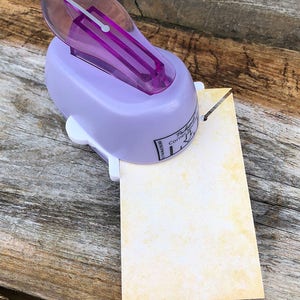 Pode incluir: Um perfurador de cantos roxo claro com pega roxa transl&uacute;cida e base branca, perfurando o canto de um papel retangular de cor esbranqui&ccedil;ada com textura amarela manchada. A ferramenta apresenta o texto impresso Corner Notch.