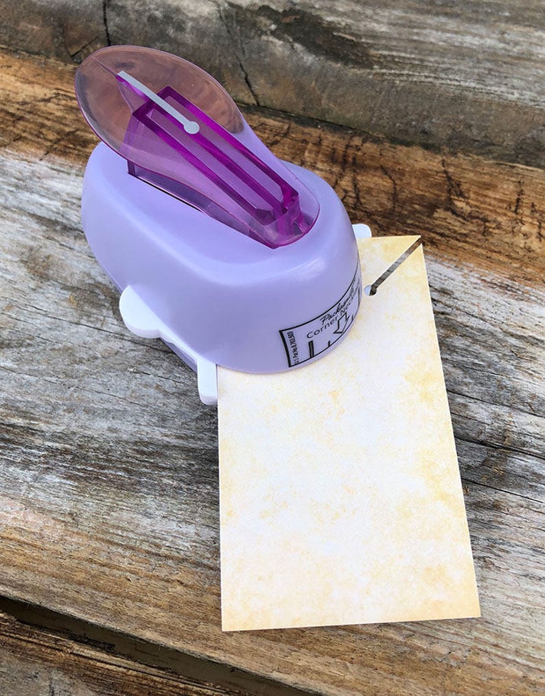 Pode incluir: Um perfurador de cantos roxo claro com pega roxa transl&uacute;cida e base branca, perfurando o canto de um papel retangular de cor esbranqui&ccedil;ada com textura amarela manchada. A ferramenta apresenta o texto impresso Corner Notch.