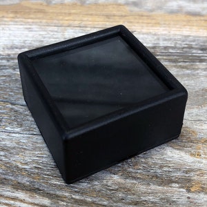 May include: A black square display case with a clear glass top. The case is made of plastic and has a smooth, matte finish.