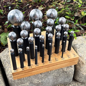May include: A collection of metal ball-end punches of varying sizes, arranged in a wooden holder. The punches have black handles and silver ball tips, with measurements in inches labeled on the wood.