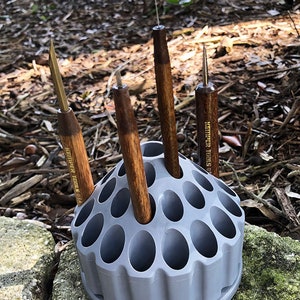 May include: A gray plastic Kemper Tools USA tool holder with several tools inserted. The tools have wooden handles and metal tips. The holder is sitting on a rock, with a blurred natural background.