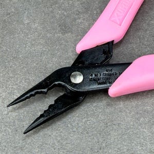 May include: Black metal pliers with pink rubberized handles. The tool features a pointed tip, serrated gripping jaws, and a central pivot point. Embossed text on the black metal body reads 494, 4 IN 1 CRIMPER PLIER, XURON CORP, USA, MADE IN USA.