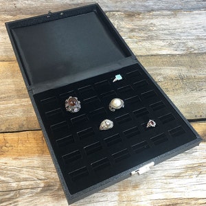 May include: Black ring box with a velvet interior and multiple compartments for storing rings. The box has a latch closure and is open to reveal several rings with different gemstones.