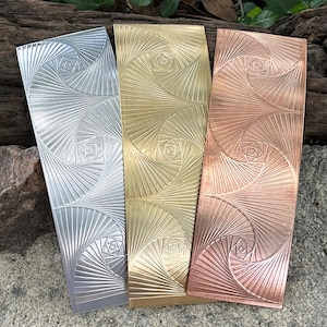 May include: Three metal bookmarks in silver, gold, and copper. Each bookmark features a spiral design with a central square pattern. The bookmarks are rectangular and have a textured surface, showcasing a geometric aesthetic.