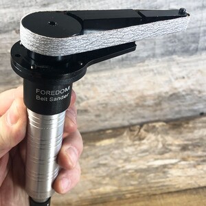 May include: A silver and black handheld belt sander with a gray sanding belt. The sander has the text "FOREDOM Belt Sander" printed on the side.