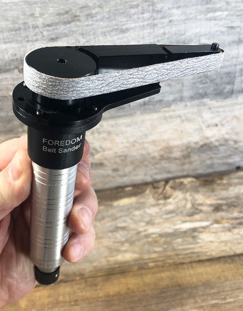 May include: A silver and black handheld belt sander with a gray sanding belt. The sander has the text "FOREDOM Belt Sander" printed on the side.