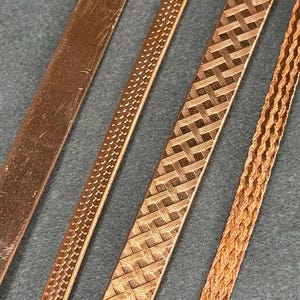May include: Close-up of four copper strips with varying textures. The first strip is smooth, the second has a dotted pattern, the third features a woven design, and the fourth is a braided style. The strips are arranged diagonally against a gray background.