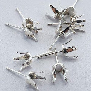 May include: A collection of six silver-toned metal earring settings, each featuring a four-prong basket design intended for holding a gemstone. These earring posts are arranged in a cluster, showcasing the metallic finish and prong structure.