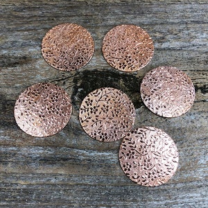 May include: Six copper-colored metal discs with a floral pattern embossed on the surface. The discs are arranged in a circle on a wooden surface.