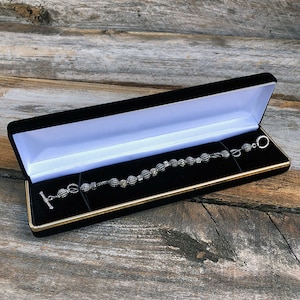 May include: A silver bracelet with black and white patterned beads in a black velvet jewelry box with a gold trim.