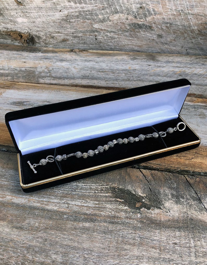 May include: A silver bracelet with black and white patterned beads in a black velvet jewelry box with a gold trim.