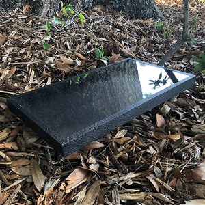 May include: A black rectangular tray with a textured surface and a reflective finish. The tray is lying on a bed of brown leaves and twigs.