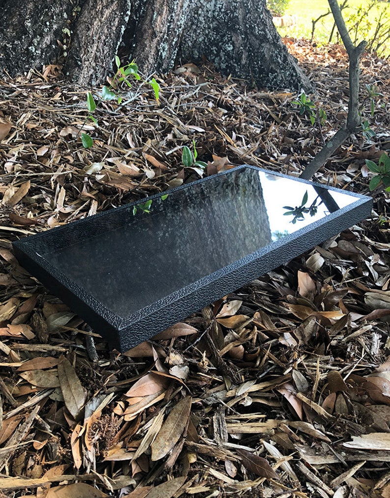 May include: A black rectangular tray with a textured surface and a reflective finish. The tray is lying on a bed of brown leaves and twigs.
