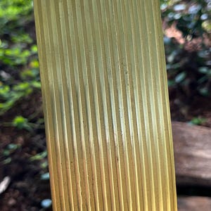 May include: A rectangular, gold-colored metal object with vertical, ridged detailing. The object is held at an angle, with a blurred green background of foliage and wood.