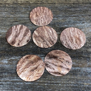 May include: Six round copper coasters with a textured surface. The coasters are arranged in a pyramid shape on a wooden surface.