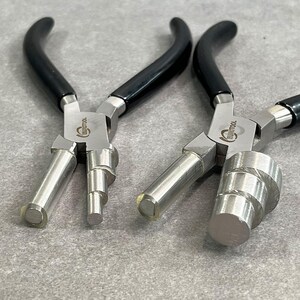 May include: Two silver metal jewelry pliers with black rubber handles. The pliers have different sized round tips for making loops in wire.