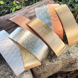 May include: A collection of seven rectangular metal strips featuring various embossed patterns including floral scrolls, geometric diamonds, wood grain textures, and leaf motifs in silver, gold, and copper tones arranged in an overlapping display.