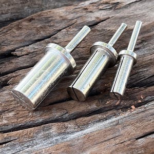 May include: Three silver metal tools with cylindrical bodies and long, thin handles are arranged on a weathered wooden surface. The tools vary in size, with a slotted design on the body. The background is a natural wood grain.