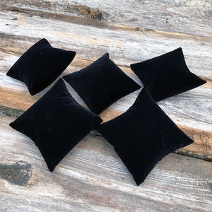 May include: Five black velvet ring display pillows. The pillows are square shaped and are arranged in a staggered pattern on a wooden surface.