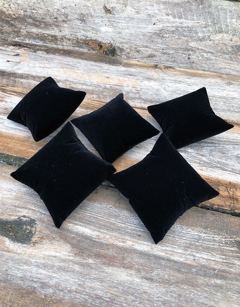 May include: Five black velvet ring display pillows. The pillows are square shaped and are arranged in a staggered pattern on a wooden surface.