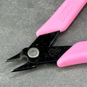 May include: A pair of Xuron brand side cutters featuring black metal blades and bright pink plastic handles. The tool has a central silver pivot pin and the brand name Xuron is embossed on one of the pink handles near the metal joint.