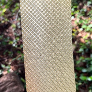 May include: A rectangular, golden-colored object with a repeating diamond pattern. The surface has a textured appearance, and the object is held against a blurred green background of foliage.
