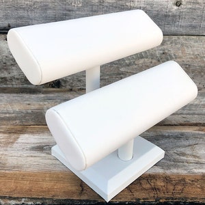 May include: White two-tiered jewelry display stand with a square base and rounded padded arms for holding bracelets or bangles.
