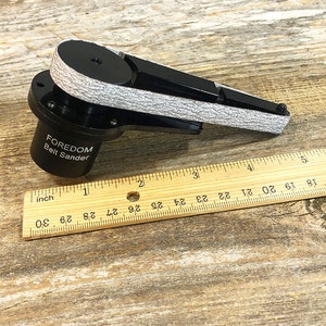 May include: A black Foredom belt sander with a gray sanding belt. The sander is about 5 inches long and is sitting on a wooden surface.