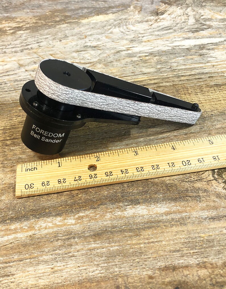 May include: A black Foredom belt sander with a gray sanding belt. The sander is about 5 inches long and is sitting on a wooden surface.