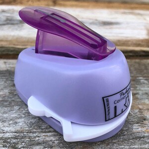 Pode incluir: Um perfurador de cantos para papel em tom lil&aacute;s claro com uma alavanca superior em pl&aacute;stico violeta escuro transl&uacute;cido. A lateral do dispositivo apresenta uma etiqueta branca com o texto preto "Corner Notch" e "U.S. Pat. No. 8,763,503".