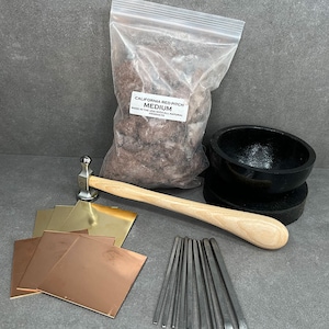 May include: A collection of jewelry-making tools and supplies, including a bag of California Red Pitch, a hammer, metal squares, a bowl, and a set of metal punches. The metal squares are copper and brass. The hammer has a wooden handle.