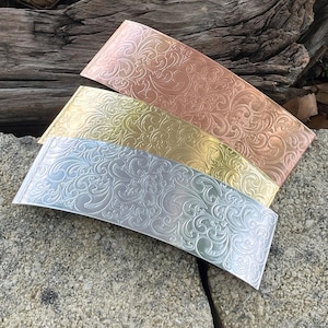 May include: Three metal hair barrettes in copper, gold, and silver, each with an ornate, swirling floral pattern. The barrettes are rectangular with a curved top edge, resting on a stone surface.