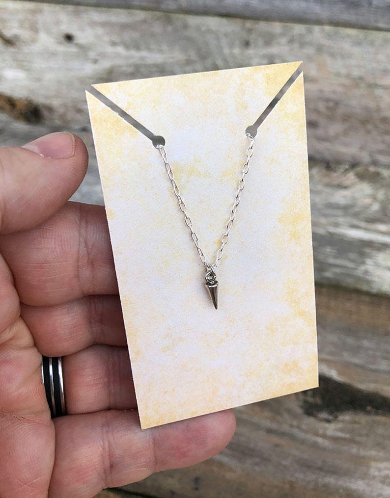 Pode incluir: Um colar de tom prateado com uma corrente delicada e um pequeno pendente em forma de cone pontiagudo. O colar &eacute; exibido num cart&atilde;o retangular amarelo-claro com textura manchada, que possui duas pequenas ranhuras no topo para a corrente.