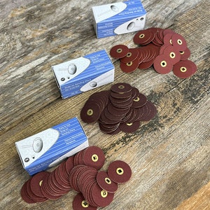 May include: Three boxes of brown sanding discs with gold centers. The boxes are blue and white and have the text "Easy to Use Snap-on Sanding Discs Discos Abrochables Abrasivos" printed on them. The sanding discs are stacked in three piles on a wooden surface.