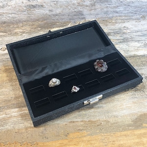 May include: Black ring box with a velvet interior and multiple slots for storing rings. Three rings are displayed in the box, including a silver ring with a large clear stone, a silver ring with a red stone, and a silver ring with a smaller clear stone.