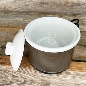 May include: A small, gray ceramic crock pot with a white lid. The crock pot has a white rim and a black handle.