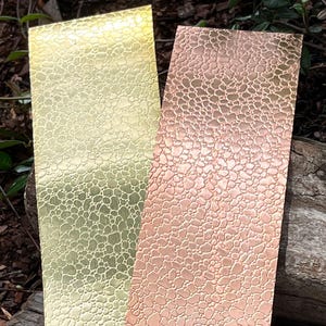 May include: Two rectangular metal sheets with a textured, organic pattern. One sheet is a bright gold color, and the other is a warm copper-rose hue. The sheets are displayed against a natural, outdoor background, suggesting a craft or design context.