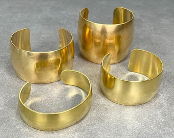 May include: Four gold-toned metal bracelets of varying widths. The bracelets are stacked on a gray surface.