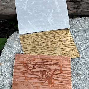 May include: Three rectangular decorative panels with bamboo leaf designs. The top panel is silver, the middle is gold, and the bottom is copper. The panels are arranged on a gray stone surface with a wooden background.