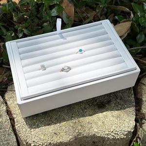 May include: A rectangular white jewelry display tray featuring multiple padded slots for organizing rings. The tray includes a small white fabric pull tab and contains three rings: one with a turquoise stone, one with a clear stone, and one with a textured band.