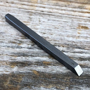 Puede incluir: Un cincel de metal plateado con una punta cuadrada. El cincel está sobre una superficie de madera.