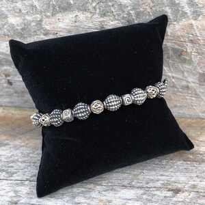 May include: A silver and gold beaded bracelet with a textured design. The bracelet is displayed on a black velvet cushion.