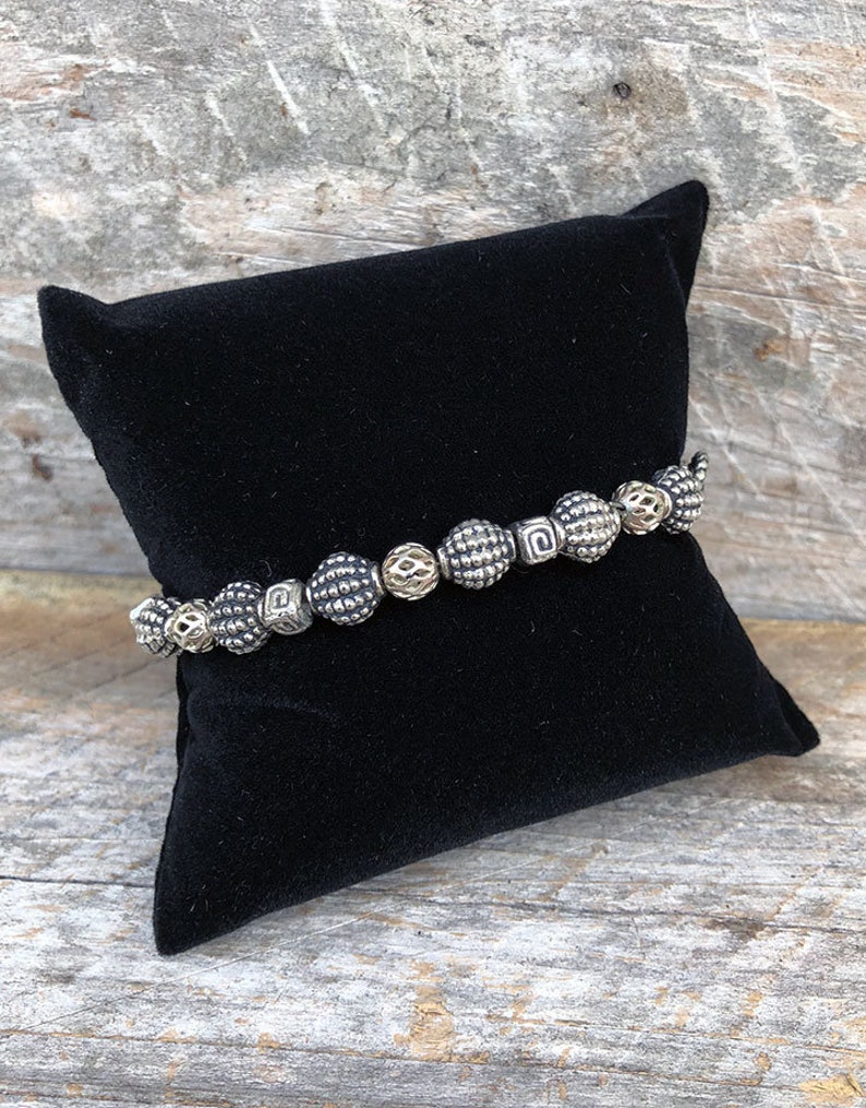 May include: A silver and gold beaded bracelet with a textured design. The bracelet is displayed on a black velvet cushion.