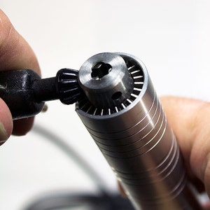 Puede incluir: Primer plano de una herramienta rotativa plateada con un portabrocas negro. La herramienta se sostiene en una mano y el portabrocas se est&aacute; girando.