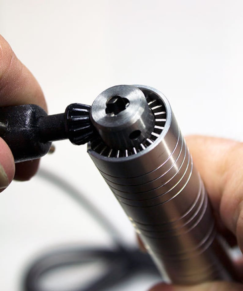 Puede incluir: Primer plano de una herramienta rotativa plateada con un portabrocas negro. La herramienta se sostiene en una mano y el portabrocas se est&aacute; girando.