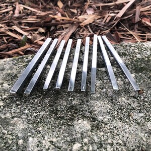 May include: Ten silver metal skewers, arranged in a row on a gray stone surface. The skewers are all the same length and width, and they are all pointed at one end.