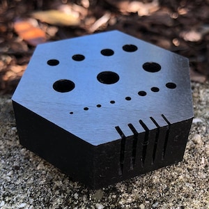 May include: A black, hexagonal wooden block with various sized holes and slots. The top surface features several circular holes of varying diameters. The side has a series of evenly spaced vertical slots. The block is resting on a stone surface.