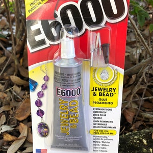 May include: A silver tube of E6000 Jewelry & Bead glue with a white cap. The label is red, white, and black with the text "E6000 Jewelry & Bead Glue" and "Permanent Bond Waterproof Dries Clear Flexible". The tube is 1.0 fl oz (29.5 ml).