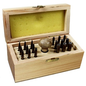May include: A wooden box containing a set of 20 metal hole punches in various sizes, ranging from 0.75 inches to 2.75 inches. The punches are arranged in a grid pattern inside the box, with a wooden hammer in the center.